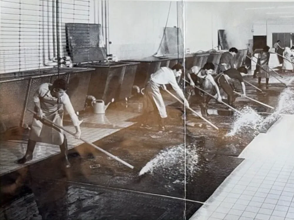 Historisches Schwarzweißfoto: Mehrere Männer in Schürzen und Gummistiefeln schrubben in einer großen gefliesten Waschhalle nasse Teppiche mit langen Bürsten, Wasser spritzt auf