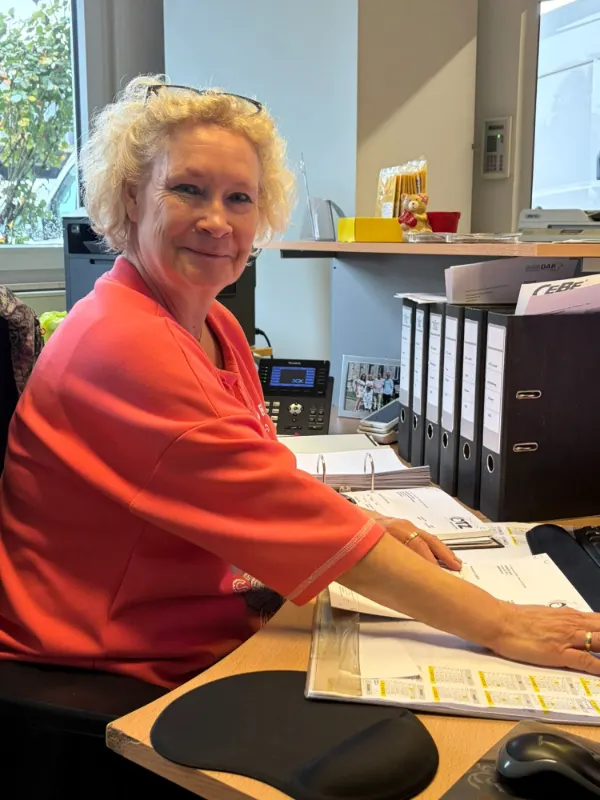Frau Windhuis sitzt lächelnd an ihrem Schreibtisch im Büro, vor ihr Ordner, Auftragsunterlagen und ein Telefon