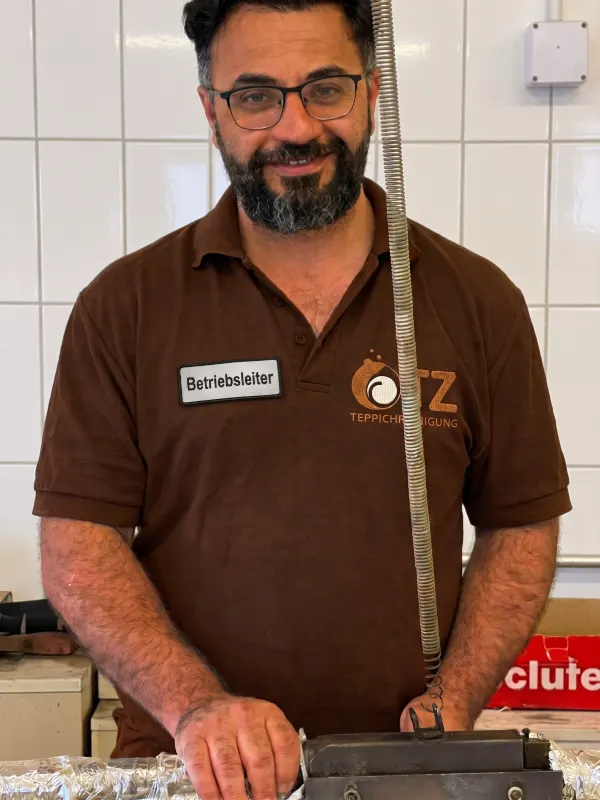 Betriebsleiter Tamim Stanikzay im braunen OTZ-Poloshirt mit Namensschild Betriebsleiter steht lächelnd an einer Maschine in der Werkstatt