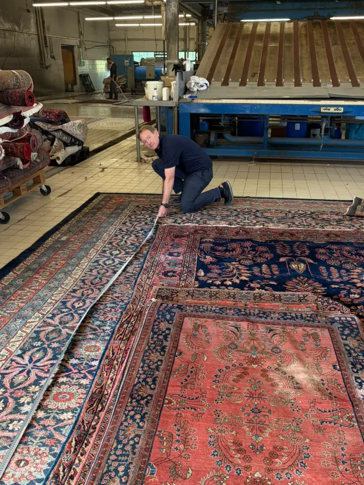 Inhaber Armin Brück legt ein langes Maßband an einen großen Orientteppich mit blauer Bordüre an, im Hintergrund die Waschhalle mit Waschanlage