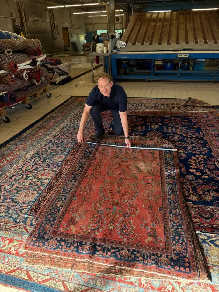 Inhaber Armin Brück kniet auf mehreren ausgebreiteten Orientteppichen und misst einen roten Teppich mit einem Maßband aus, links stehen gerollte Teppiche auf einem Wagen